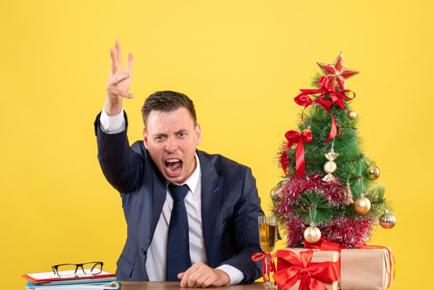 商务愤怒的男人坐在圣诞树旁大声喊叫 黄色墙上挂着礼物行政人而