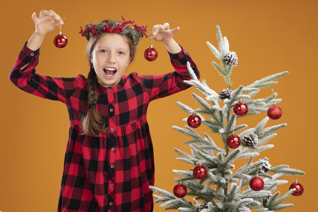 站着小女孩穿着格子衬衫 戴着圣诞花圈 手里拿着圣诞球 看着摄像机 站在橙色背景下的圣诞树旁 开心而天真地微笑着女孩格子衬衫