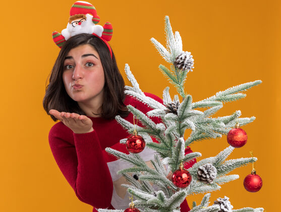 站着穿着圣诞毛衣 戴着滑稽头带 站在橙色背景下的圣诞树旁对着摄像机吹着吻的快乐小女孩年轻女孩搞笑