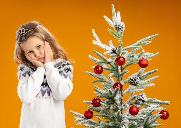花环悲伤的歪着头的小女孩站在圣诞树旁 戴着头饰 脖子上戴着花环 手放在脸颊上 橘黄色的背景隔离开来放圣诞节头