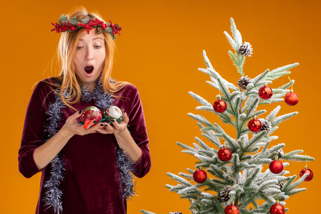看惊讶的年轻漂亮女孩站在圣诞树旁边 穿着红色的裙子 脖子上戴着花环 抱着圣诞球 看着橙色背景下的圣诞球脖子圣诞节惊讶