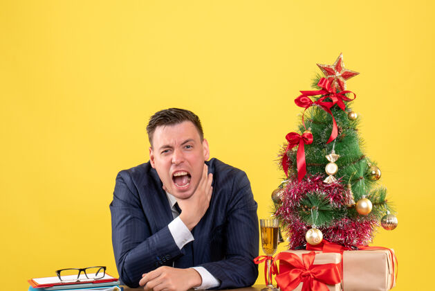 商人前视图愤怒的男子勒死自己的手坐在桌旁圣诞树和礼物上的黄色墙壁桌子生意坐着