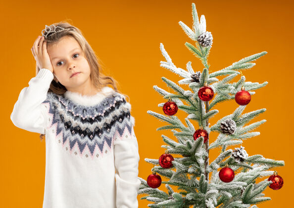 花环疲惫的歪着头的小女孩站在圣诞树旁 戴着头饰 脖子上戴着花环 手放在头上 孤立在橙色背景上树站倾斜