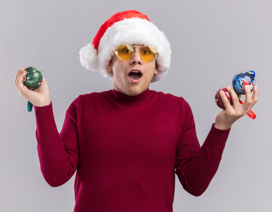 球惊讶的年轻人戴着圣诞帽 戴着眼镜 手里拿着圣诞树上的球 背景是白色的圣诞节惊讶帽子