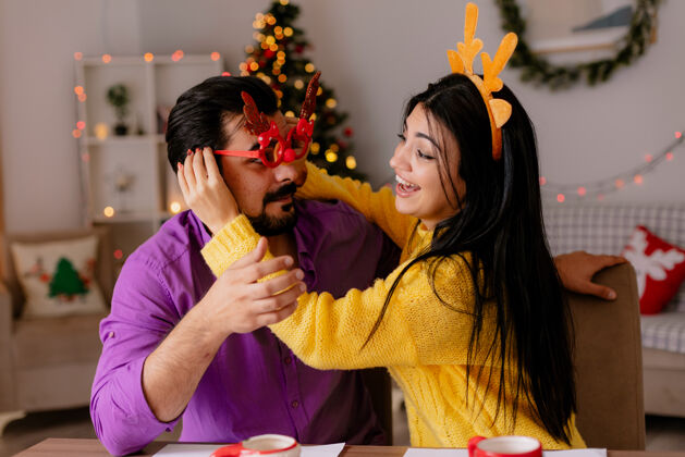 圣诞一对年轻漂亮的情侣坐在桌子旁 拿着饼干 一起开心 在圣诞装饰的房间里 以圣诞树为背景饼干桌子有