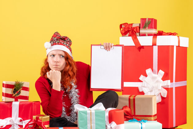 文件坐在圣诞礼物周围的年轻女子的正面视图 黄色墙上有文件说明前面礼物礼物