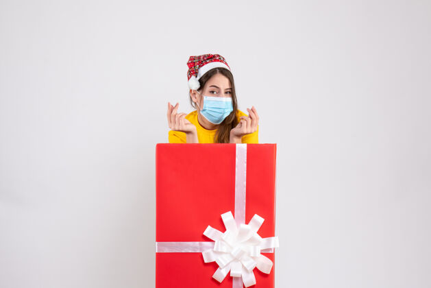 庆祝戴着圣诞帽和面具的小女孩站在白色圣诞礼物后面年轻礼物站着