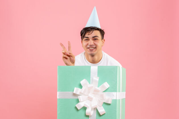年轻隐藏在礼物里的年轻人在粉红色墙上微笑的正面视图盒子包装生日