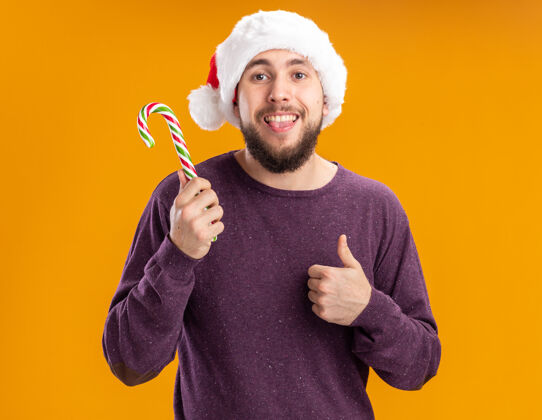 拇指快乐的年轻人穿着紫色毛衣 戴着圣诞帽 手里拿着糖杖 伸出舌头 竖起大拇指站在橙色的背景上帽子手杖坚持