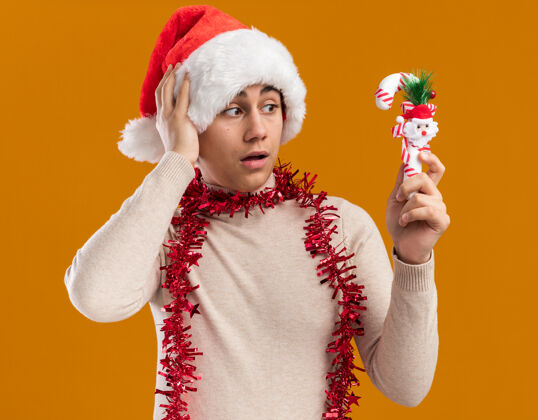 抱着想着戴着圣诞帽脖子上戴着花环的年轻人 看着黄色背景上的圣诞糖果圣诞节帽子糖果