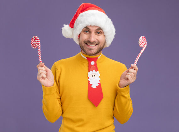 手杖微笑着的白人年轻人戴着圣诞帽 打着圣诞领带 手里拿着圣诞甜手杖 看着紫色背景上孤立的摄像机相机年轻领带