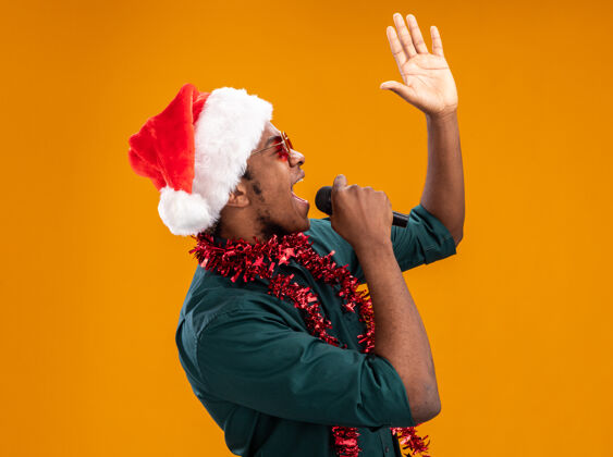 胳膊戴着圣诞帽 戴着花环 戴着眼镜的非裔美国人高举着手臂站在橙色背景上对着麦克风喊叫美国人眼镜举起来