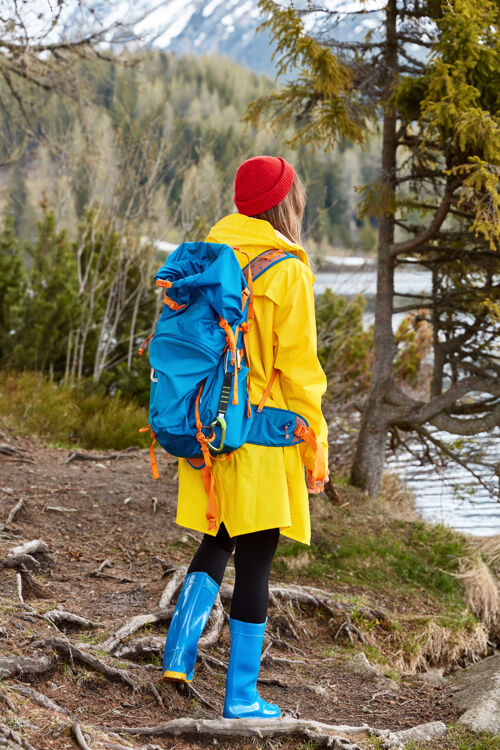 欣赏背着背包的徒步妇女站在湖边 欣赏大自然的景色 穿着黄色雨衣和胶靴山享受风景