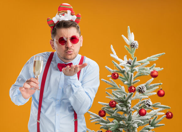 吊带戴着吊带领结的年轻人站在圣诞树旁 戴着圣诞老人和红眼镜 手里拿着一杯香槟 欢快地在橙色的背景上吹着一吻弓圣诞树边缘