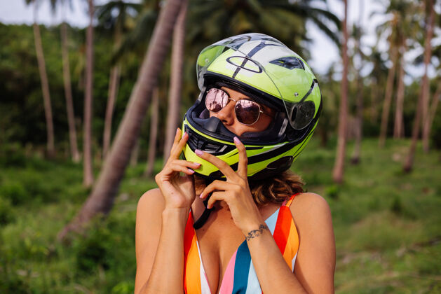 光明在棕榈树下热带田野的丛林中 穿着黄绿色摩托车头盔和彩色浅夏装的美丽骑手妇女的肖像幸福热带骑自行车