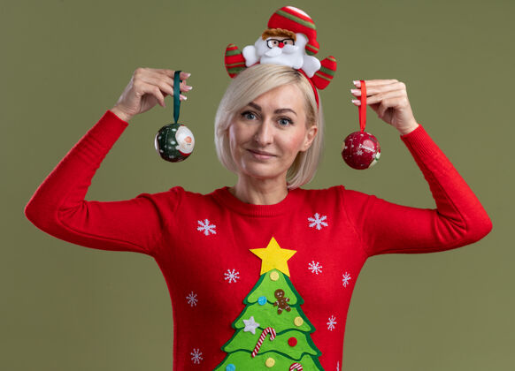 拿着高兴的中年金发女人戴着圣诞老人的头带和圣诞毛衣拿着圣诞饰品近头看相机隔离在橄榄绿的背景看相机圣诞老人