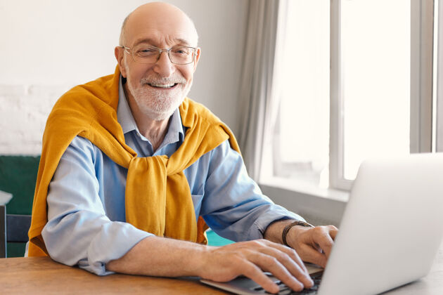 成熟开朗成熟的胡须秃顶的男性企业家戴着眼镜和毛衣 穿着蓝色的正式衬衫 一边开心地微笑 一边在便携式电脑上输入键盘 一边在午休时间玩电子游戏电脑浏览帅哥