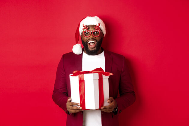礼物圣诞节英俊的非洲裔美国人戴着派对眼镜 戴着圣诞帽 手里拿着新年礼物年轻人男孩时尚