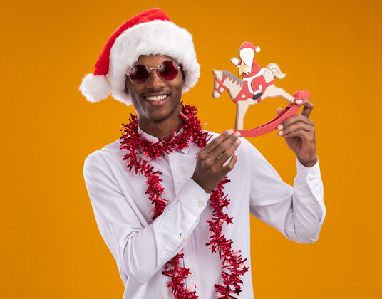 快乐快乐的非洲裔美国年轻人戴着圣诞帽 戴着眼镜 脖子上戴着金箔花环 抱着摇摇木马上的圣诞老人 看着橙色背景上孤立的摄像机雕像金属片脖子