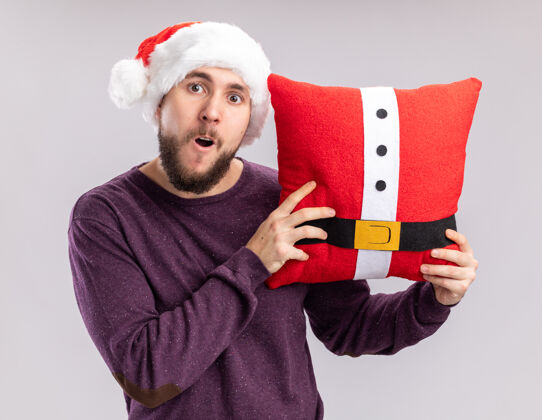 帽子穿着紫色毛衣 戴着圣诞帽的年轻人抱着滑稽的枕头 站在白色的背景下惊讶地看着镜头毛衣搞笑惊讶