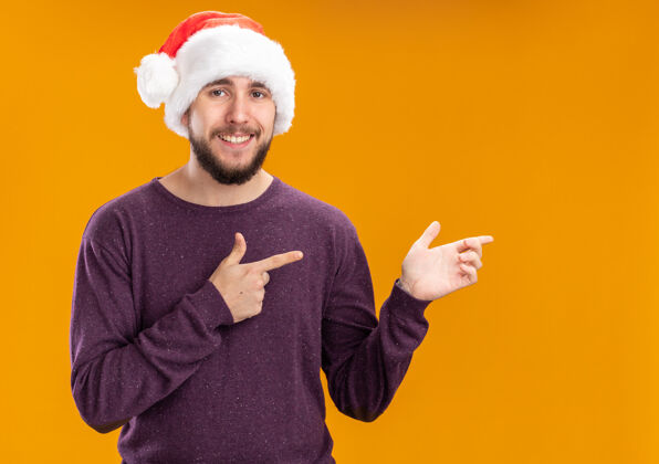 站在一边身穿紫色毛衣 戴圣诞帽的年轻人看着摄像机 兴高采烈地微笑着 用食指指着站在橙色背景上的一边食指指指点点毛衣