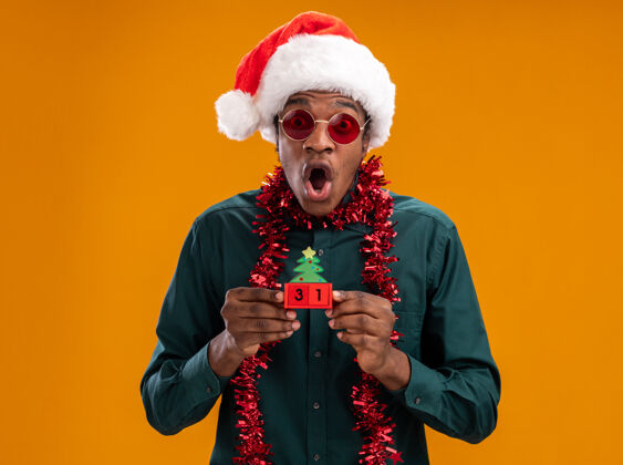 花环非洲裔美国人戴着圣诞帽 戴着花环 戴着太阳镜 拿着新年约会的玩具立方体 站在橙色的背景下 看着镜头 既惊讶又震惊年太阳镜日期