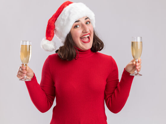 毛衣穿着红色毛衣 戴着圣诞老人帽的年轻女孩举着两杯香槟看着镜头站在白色的背景下开心而愉快地微笑着女孩香槟站着