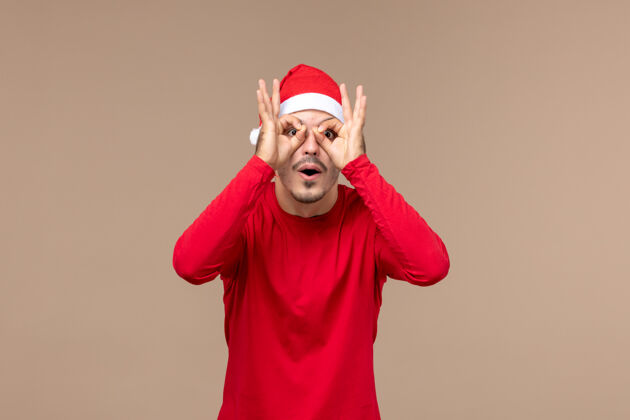 男人前视图年轻的男性透过手指看棕色背景的情感圣诞假期看表情通过