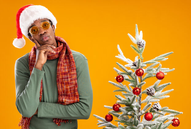 脖子戴着圣诞帽 脖子上围着围巾的非裔美国人站在橙色背景下的圣诞树旁 带着怀疑的表情看着摄像机围着怀疑圣诞老人