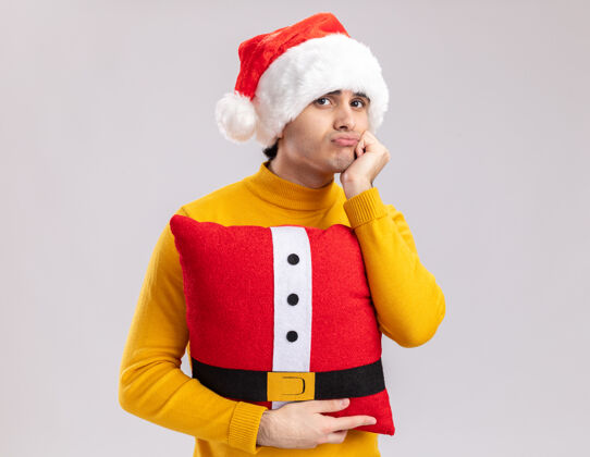 无聊穿着黄色高领毛衣 戴着圣诞帽的年轻人抱着圣诞枕头 站在白色背景下 看着相机 又累又无聊枕头年轻圣诞老人