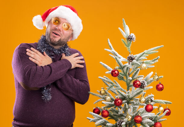 附近令人印象深刻的中年男子戴着圣诞帽 脖子上戴着金箔花环 戴着眼镜 站在装饰好的圣诞树旁 抬起头来 双手交叉放在橙色的墙上保持周围圣诞老人