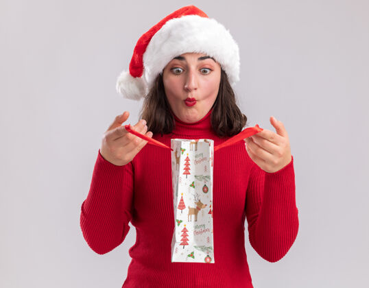 礼物穿着红色毛衣 戴圣诞帽的年轻女孩拿着彩色纸袋和圣诞礼物 打开袋子 站在白色背景下惊讶地看着里面毛衣女孩圣诞老人