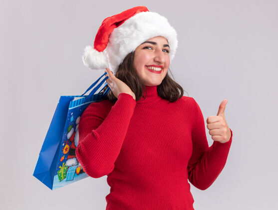 站着穿着红色毛衣 戴着圣诞帽 手里拿着彩色纸袋 手里拿着圣诞礼物 微笑着看着镜头 竖起大拇指站在白色的背景上毛衣女孩年轻人