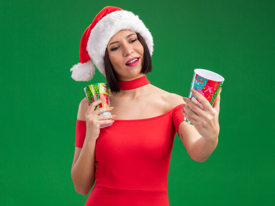 举行高兴的年轻女孩戴着圣诞帽 手里拿着塑料圣诞杯 伸着一只眼睛 看着它孤立的绿色背景伸展杯子圣诞节