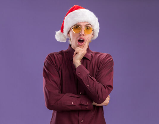 帽子令人印象深刻的年轻金发男子戴着圣诞老人的帽子和眼镜 手放在下巴上 看着紫色背景上与复制空间隔离的相机圣诞老人金发印象深刻