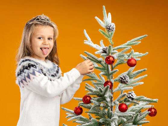 女孩高兴的小女孩站在圣诞树旁边戴着头饰 脖子上戴着花环 把玩具挂在树上孤立的橙色背景上脖子圣诞快乐花环