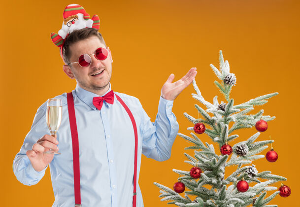 眼镜戴吊带领结的年轻人站在圣诞树旁 戴着圣诞老人和红眼镜 手里拿着一杯香槟酒 在橙色的背景下快乐而愉快玻璃杯举行轮辋