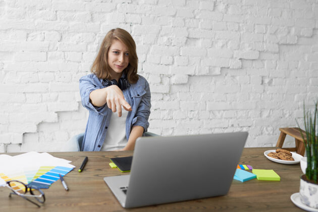 办公室肢体语言和现代科技概念情绪化的年轻女子戴着耳机 坐在打开的笔记本电脑前 微笑着用食指指着屏幕 看到一些有趣的东西女商人查看电脑