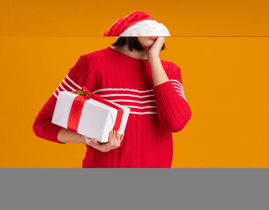 持有关心的年轻女孩戴着圣诞帽拿着礼包看着相机手放在脸上隔离在橙色背景与复制空间帽子圣诞老人关心