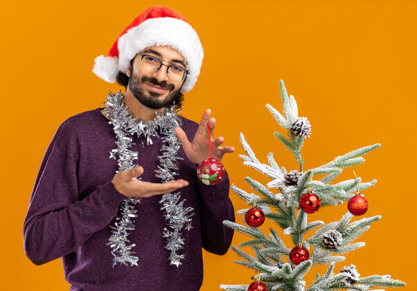 脖子站在圣诞树旁的年轻帅哥很高兴 戴着圣诞帽 脖子上戴着花环 指着橙色背景上孤立的圣诞球附近帅气站着