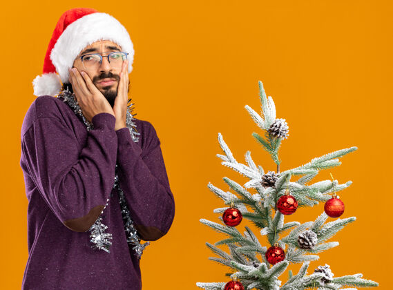 树站在圣诞树旁的悲伤的年轻帅哥戴着圣诞帽 脖子上戴着花环 手放在脸颊上 橙色背景下孤立着站着帽子圣诞节