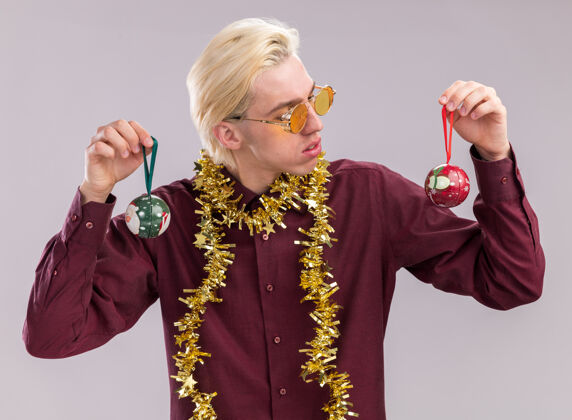 年轻的困惑的年轻金发男子戴着眼镜 脖子上戴着金属丝花环 手里拿着圣诞饰品 看着一个孤立的白色背景圣诞饰品周围拿着