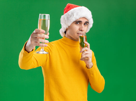 站着一个穿着黄色高领毛衣 戴着圣诞老人帽的年轻人 手里拿着两杯香槟 站在绿色的背景上 快乐而积极地看着镜头圣诞老人拿着帽子