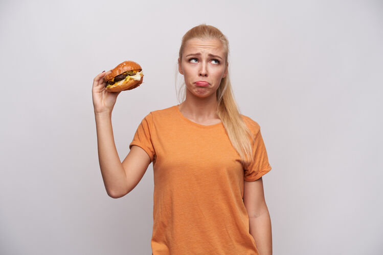 T恤心烦意乱的年轻长发金发女士身穿橙色t恤 站在白色背景下 悲伤地看着一边 皱着额头 同时举起手拿着不健康的食物汉堡饮食美丽