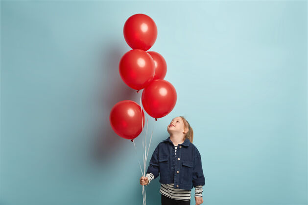 庆祝快乐的小女孩抬起头 聚精会神地看着红色的气球 穿着牛仔时髦的夹克 准备庆祝生日 在蓝色的墙壁上做模特 在室内玩耍为孩子们举办派对小快乐积极
