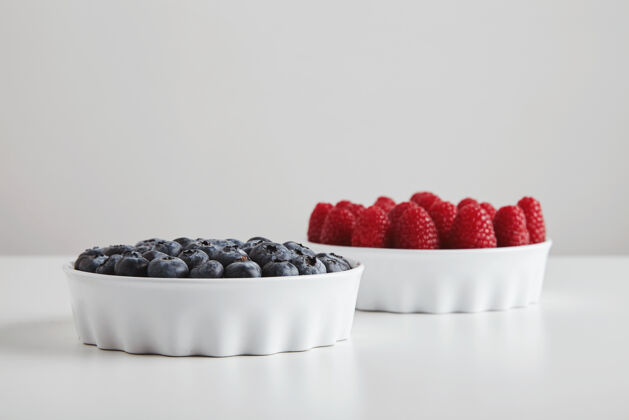 食物一堆成熟的覆盆子和蓝莓准确地放在白色桌子上的陶瓷碗里作物甜食碗