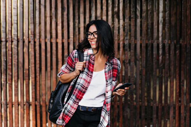 谈话穿着格子衬衫的精致黑发女孩在木墙上摆姿势带着背包的快乐拉丁女模特的户外照片快乐电话手机