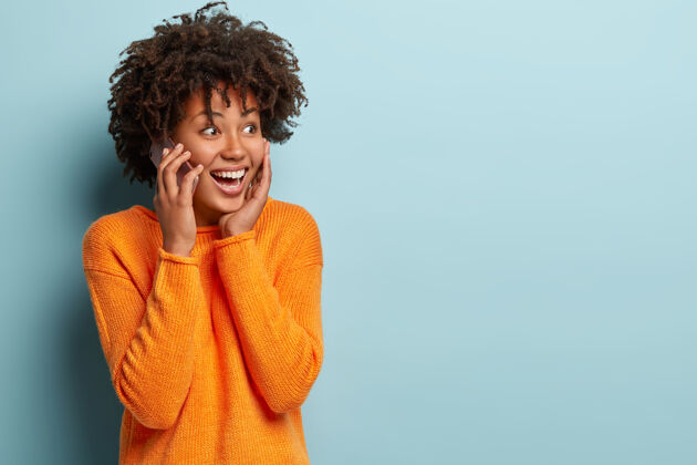 乐观情绪开朗的女性用手机交谈 与对话者分享印象 享受现代科技 穿着休闲服 隔着蓝色的墙壁 通过手机给朋友打电话设备旁白非洲