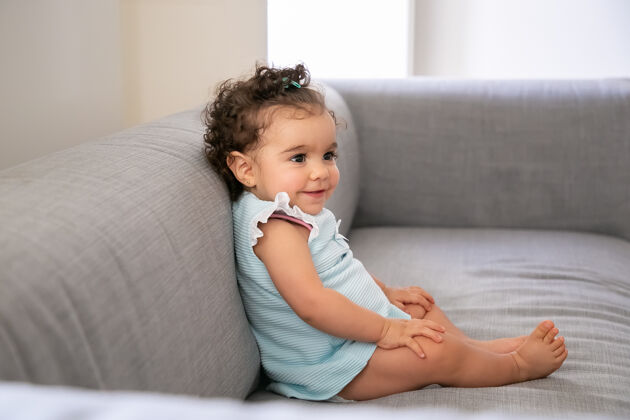 头发快乐的深色卷发女婴 穿着淡蓝色的衣服 坐在家里灰色的沙发上 微笑着看着别处家里的孩子和童年的概念童年混合成人