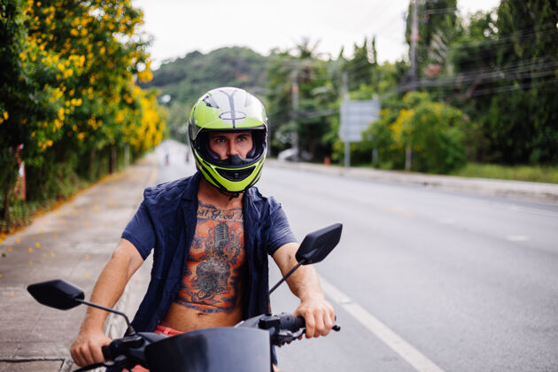 乐趣在泰国繁忙的公路边 骑摩托车的戴着黄色头盔的男性纹身车手的肖像热带比赛交通工具